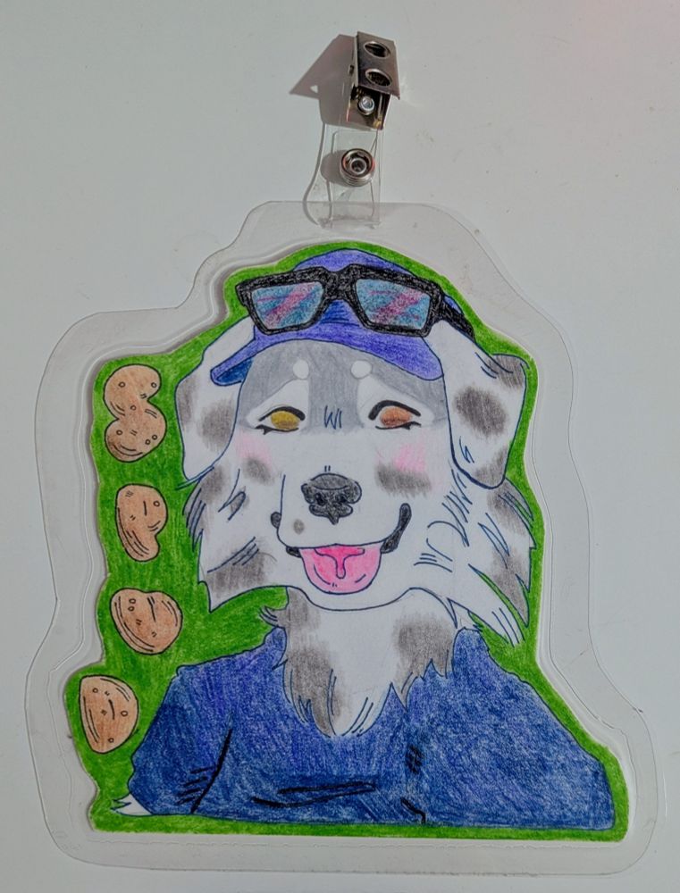 A physical badge of a dog furry in a t-shirt and baseball cap. She is looking at the viewer with soft eyes and her tongue is sticking out in a blep. To the left her name, "Spud", is spelled out with letters made out of potatoes