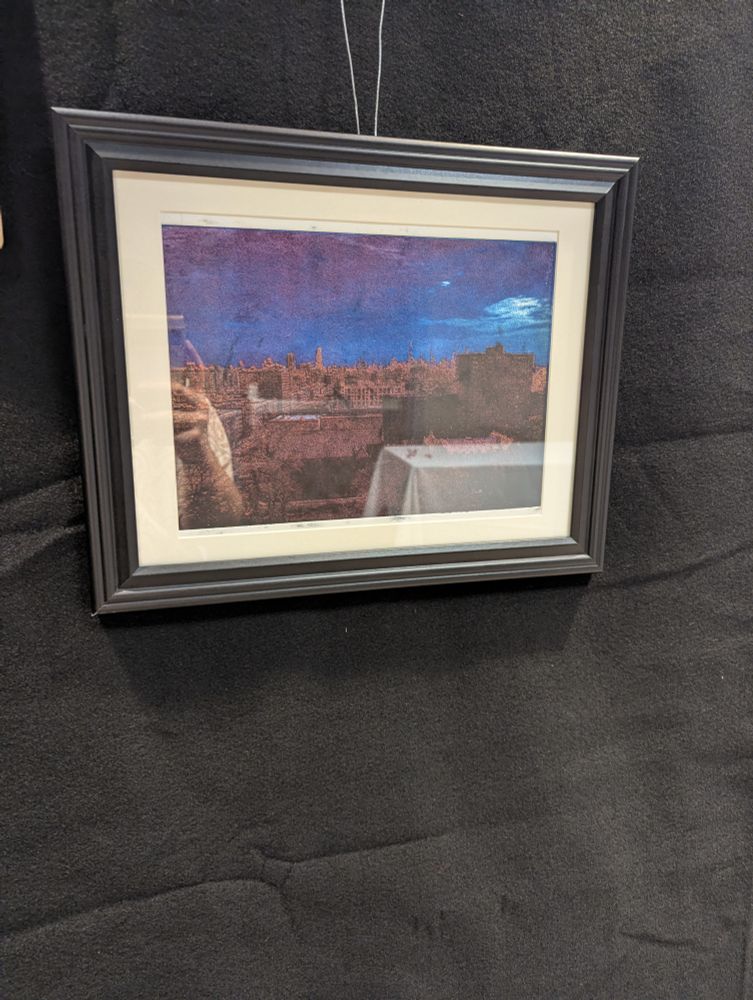 A photo of a drypoint of the Manhattan skyline, framed, hung on a black temporary wall.