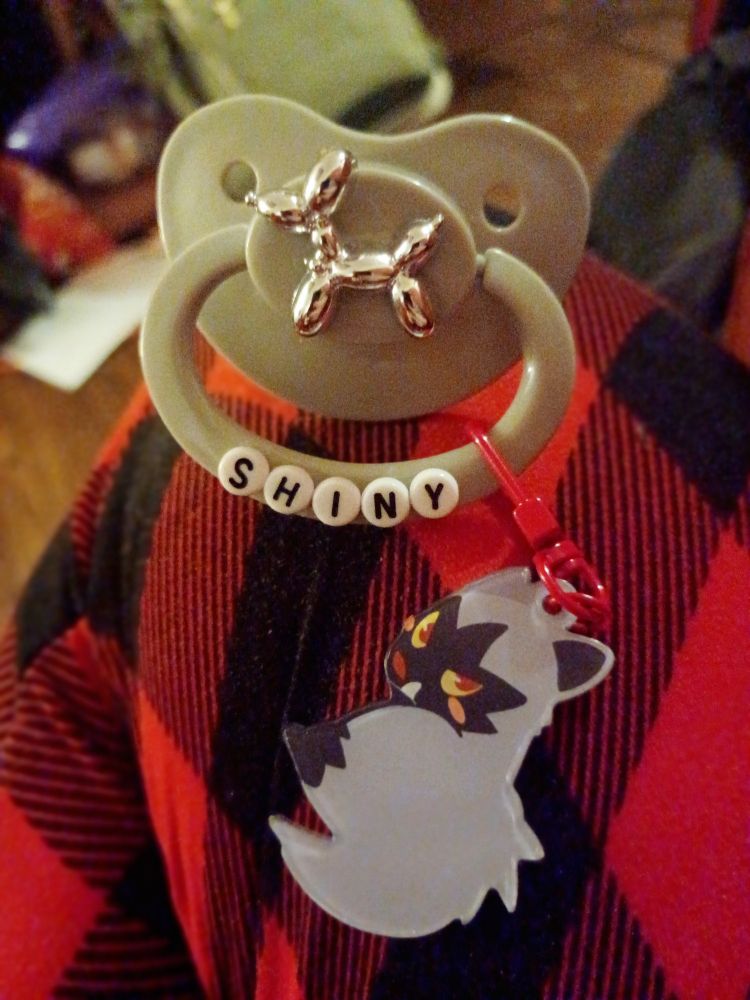 my new pacifier sits on my leg. the pacifier is grey and has a shiny chrome balloon dog on the button, and the handle says shiny in beads. an art clip of a poochyena is attached to its handle. my red plaid pants match the red of the clip.