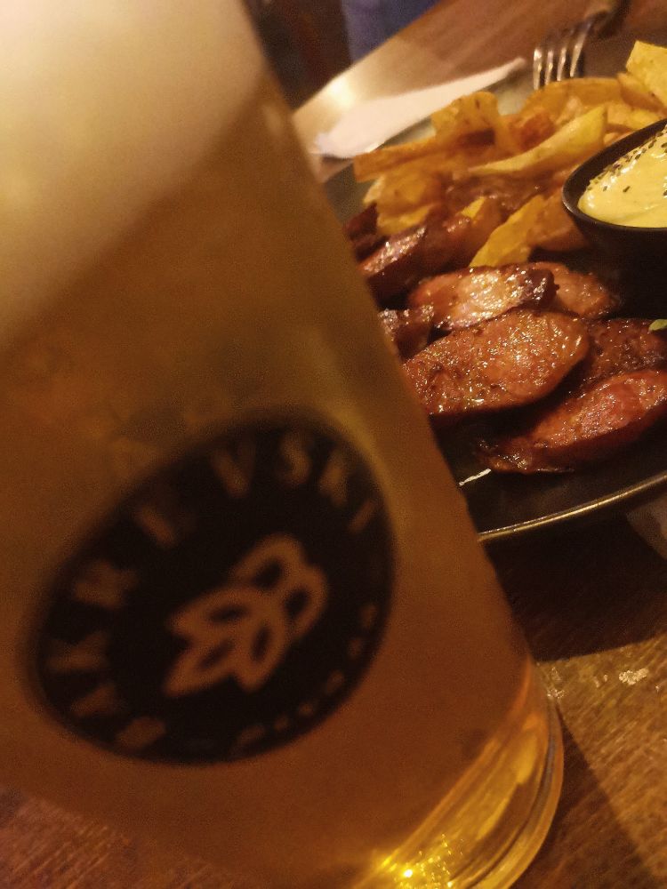 ENG:  Wooden table featuring a frothy glass of beer and a plate of food. The plate contains sliced sausages, French fries, and a small bowl of dipping sauce.

МК: Фотографија на дрвена маса со чаша пиво со пена и чинија со храна. Во чинијата има сечкани колбаси, помфрит и мала чинија со сос.