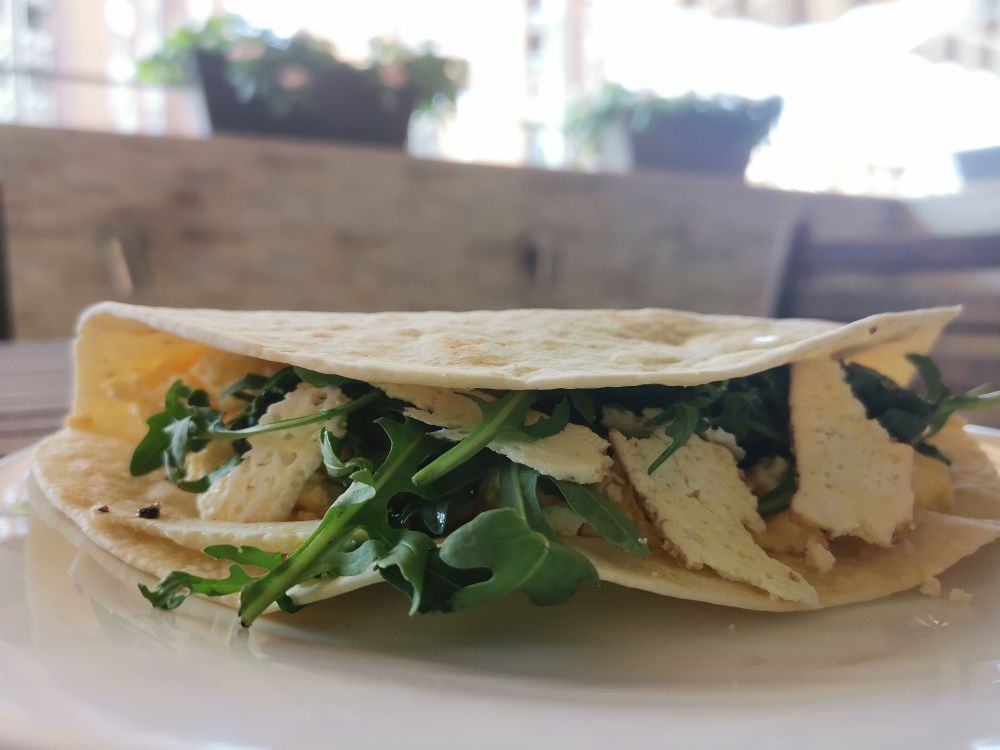 EN:
Close-up of a folded tortilla wrap on a white plate, filled with arugula, olive oil, slices of goat cheese, and scrambled eggs with dry chilly and quark.

MK:  
Свиткана тортиља на бела чинија, полнета со рукола, маслиново масло, парчиња козјо сирење и кајгана со сува чили пиперка и урда.