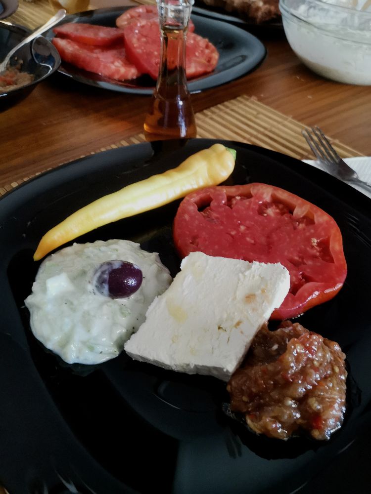 ENG: A black plate with food on a table. The plate features a yellow pepper, a slice of tomato, a piece of cheese, tarator salad with an olive, and balkan roasted pepper spread. In the background, there’s another plate with tomato slices and a rakiya.

МКД: Црна чинија со храна на маса. На чинијата има жолта лута пиперка, парче домат, парче сирење, таратор со маслинка и пинџур. Во позадина има друга чинија со парчиња домат и чоканче со ракија.