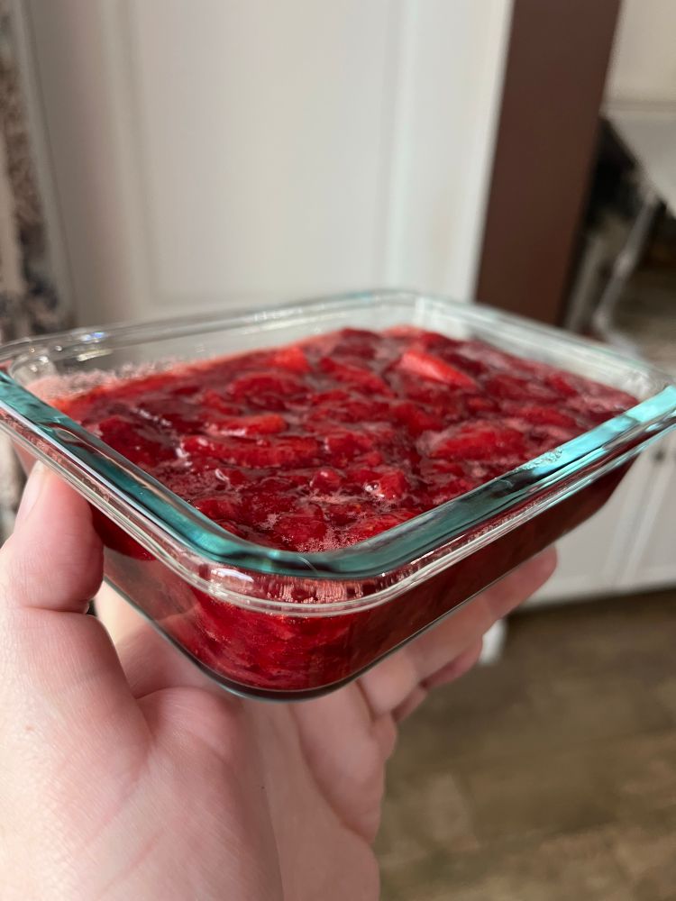 A left hand holding up one of the glass containers of strawberry sauce closer to the camera into the light