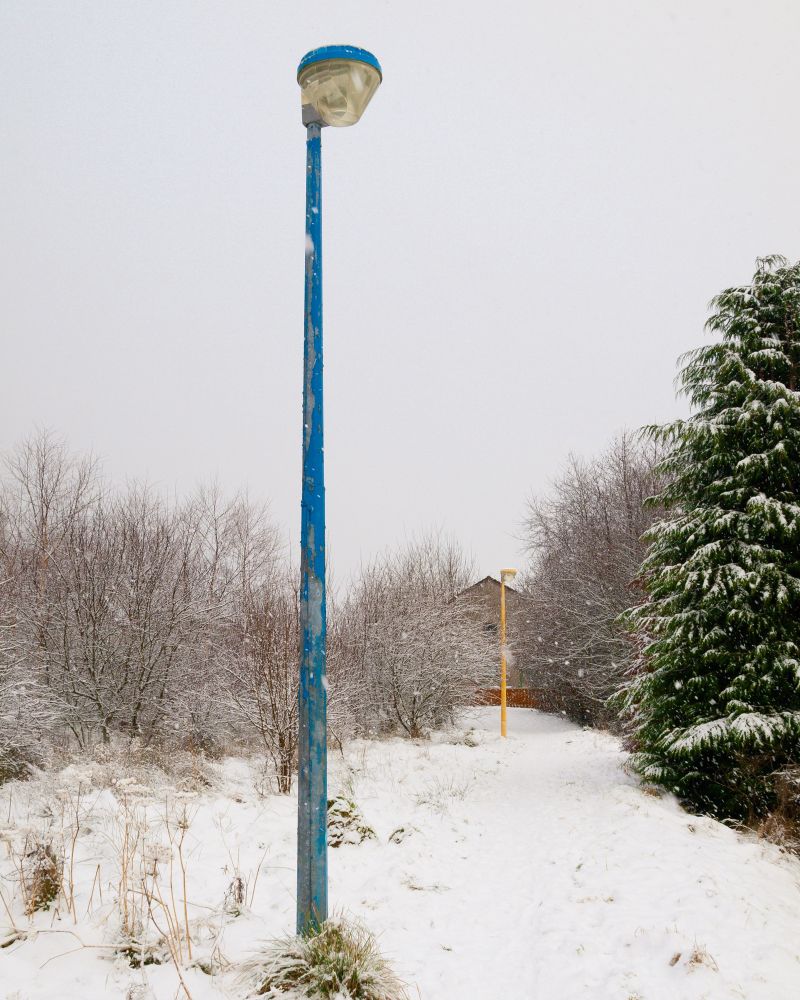 Colourful but neglected-looking lamp posts on a footpath in the snow; the nearest has cracked and peeling blue paint.