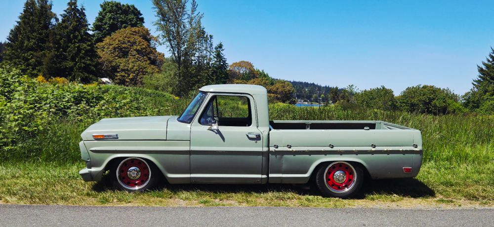 An older lowrider Ford truck in clean condition. Light green body with red hubcaps. Parked on the side of the road, with green grass and trees, and a clear blue sky as a backdrop.