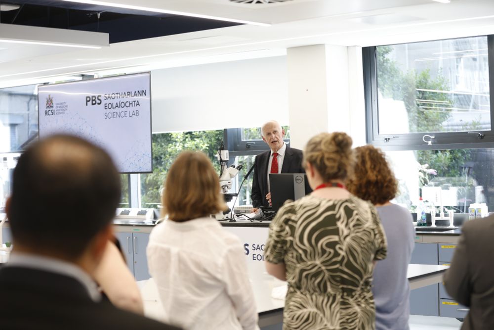 Professor Cathal Kelly, Vice Chancellor of RCSI University of Medicine and Health Sciences, addressing an audience at the opening of the redeveloped School of Pharmacy and Biomolecular Sciences science lab. He stands at a podium in a modern laboratory, with attendees listening in the foreground and a large screen displaying the lab's branding.