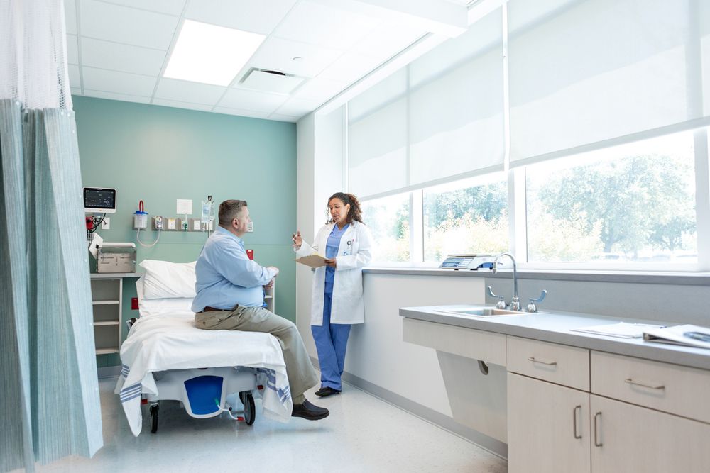 A healthcare professional speaks with a patient in a hospital room. The patient is seated on a hospital bed, and the clinician stands holding a clipboard, engaged in conversation. Sunlight streams through large windows behind them.