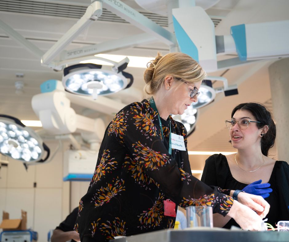 An instructor wearing a black floral blouse provides guidance to a student in a black top and gloves in a surgical simulation room with overhead medical lighting.

