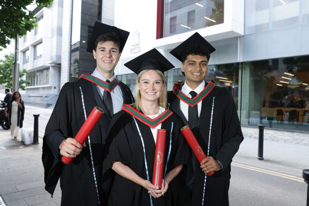 Three graduates in black gowns and mortarboards stand smiling outside the RCSI building. Each holds a red diploma scroll and wears a green and red academic hood.