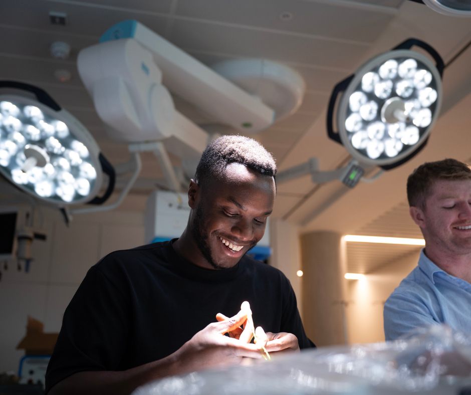 Two students, one smiling broadly, work together on a hands-on surgical or technical task in a bright lab environment with operating theatre-style lights above.
