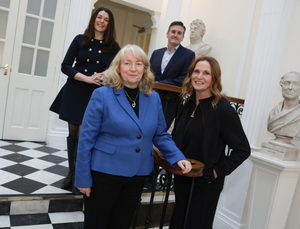 Four people pose together in a formal indoor setting with classical busts and columns in the background. Two women stand in the foreground—one in a blue blazer and black top, the other in a black outfit. Behind them, a woman in a navy dress and a man in a blue suit smile while standing on stairs. The setting has a black and white checkered floor and white walls with decorative molding.