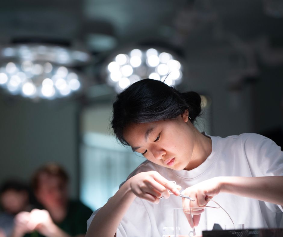 A focused student in a white top practices suturing or surgical knot-tying using forceps and thread under bright surgical lights in a clinical skills lab.