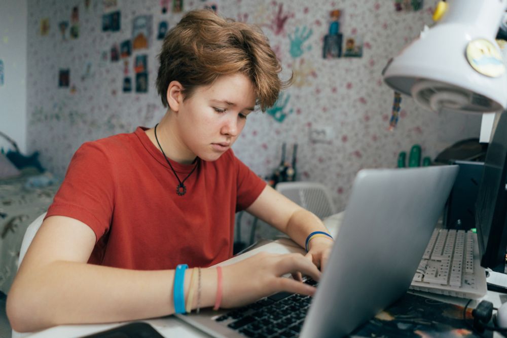 A young person wearing a red t-shirt and wristbands is focused on working on a silver laptop at a desk in a bedroom. The room is decorated with handprints and photos on the wallpapered wall behind, and a white desk lamp is visible on the right. The person appears to be deeply engaged in their task, possibly studying or working on a project.