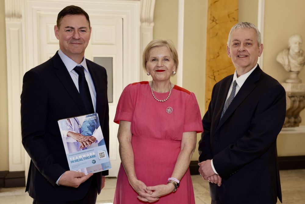 Three professionally dressed individuals stand side-by-side indoors, posing for a photo in a formal setting with classical architectural elements. The man on the left, wearing a dark suit and blue tie, holds a brochure titled "Teamwork in Healthcare." The woman in the center wears a vibrant pink dress with a pearl necklace and red lipstick, and the man on the right also wears a dark suit and tie. They are smiling, and a marble bust is visible in the background on the right.