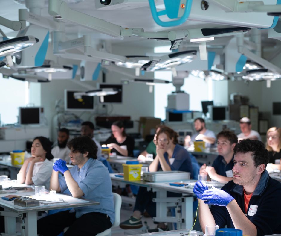 A classroom of students seated at lab benches in a surgical training facility, attentively watching an instructor or demonstration, some wearing gloves and lab badges.