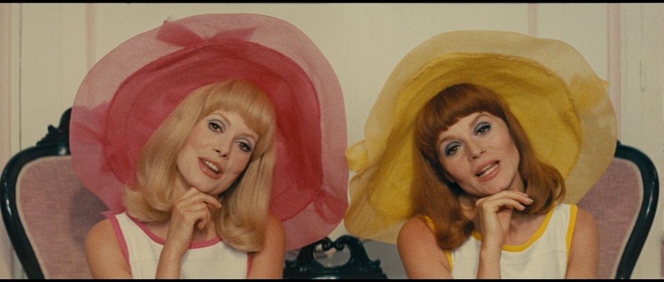 Delphine and Solange in big hats staring down the barrel of the camera, singing