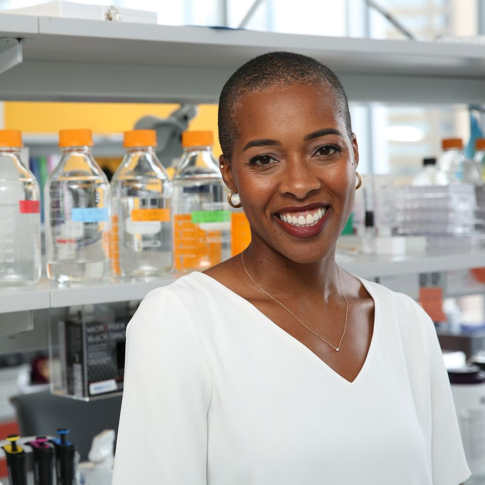 Dr. Marlin, a Black woman neuroscientist, is smiling proudly in her lab space. She’s wearing a white shirt, simple necklace, small hoop earrings, dark red lipstick and has a beautifully shaved head.