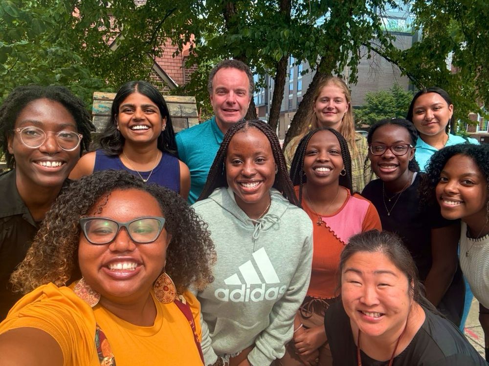 The 2025-26 cohort of incoming first year and second year MIND post-baccalaureate researchers with their program leadership. The group is smiling for a selfie outside in front of trees.