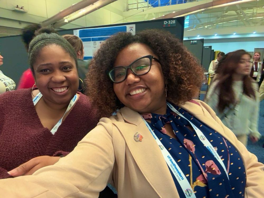 Two Black women neuroscientists smiling widely while taking a selfie. Dr. Williams on the left has her hair in a bun and is wearing a burgundy sweater. Dr. Dukes on the right has a curly Afro and is wearing a dark blue blouse with pink flowers and gold dots. She is also wearing a light pink blazer, Black In Neuro pin, and black glasses.