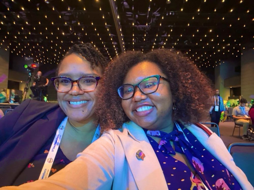 Two Black women PhDs smiling widely while taking a selfie. Dr. Watt on the left is wearing a dark blue blouse with a navy blazer and ombré colored glasses. Dr. Dukes on the right has a curly Afro and is wearing a dark blue blouse with pink flowers and gold dots. She is also wearing a light pink blazer, Black In Neuro pin, and black glasses. 