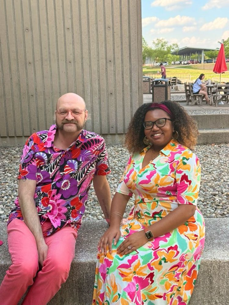 A photo of Dr. Troy Roepke and Dr. Angeline Dukes sitting outside on Rutgers campus. Troy is wearing a pink and purple floral shirt with pink pants. Angeline is wearing a cream dress with watercolor pink, orange and red flowers with green leaves.