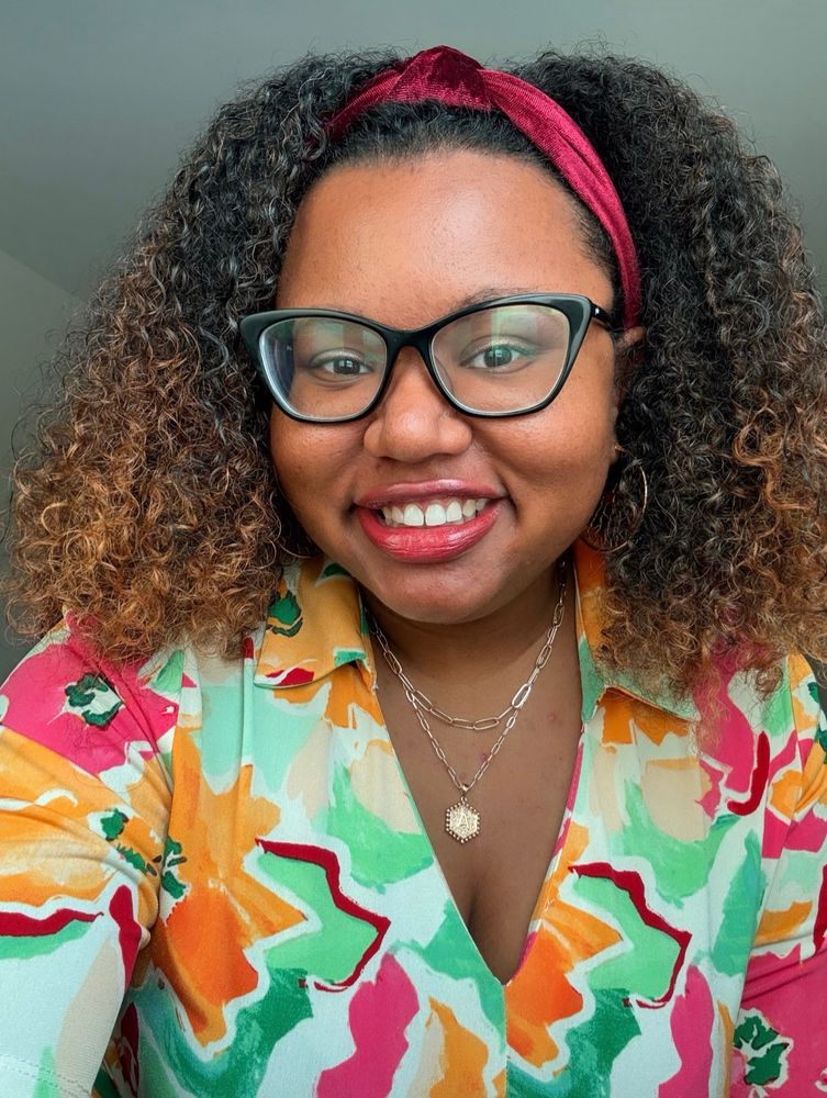 A Black woman with long curly hair taking a selfie. She is wearing a cream dress with orange and pink flowers that has green watercolor leaves, gold necklaces and a burgundy headband. She also has black glasses and red lipstick on.