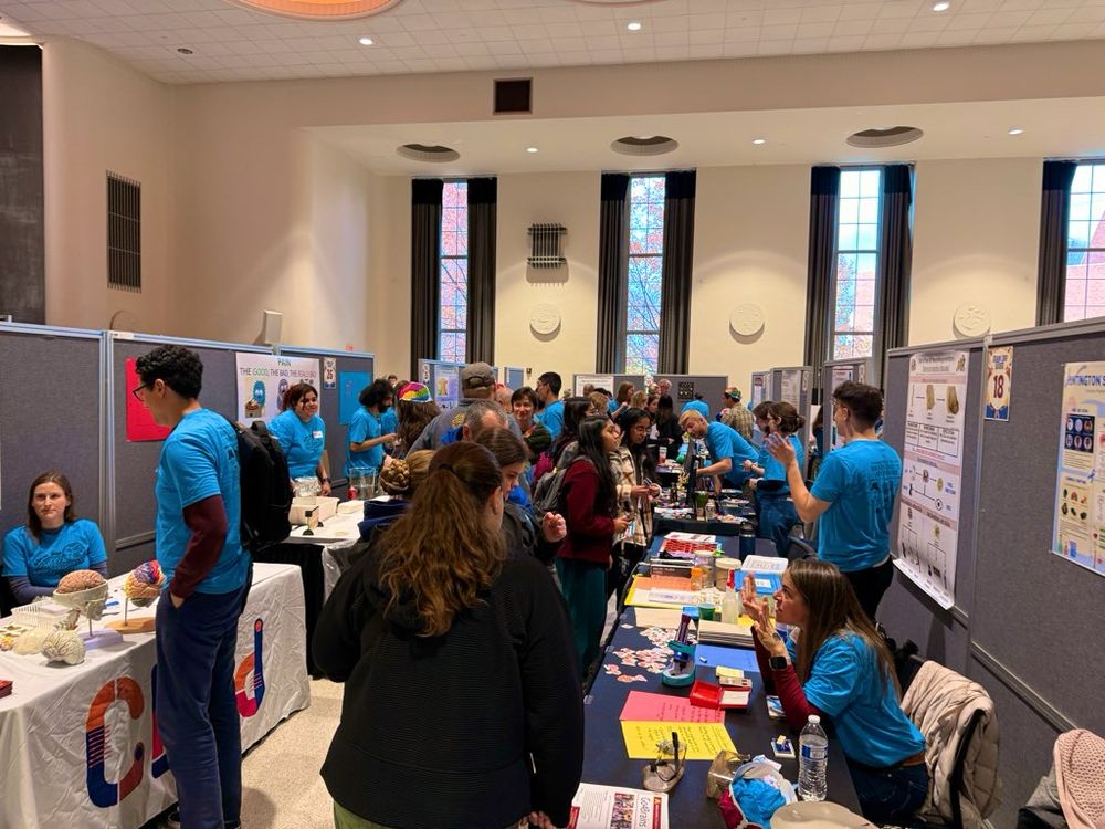 A side view down one aisle of the UMN Brain Day outreach event. Booths staffed by trainees and faculty in blue t-shirts line the aisle. Community members are actively engaged in learning about neuroscience and the work the researchers do.