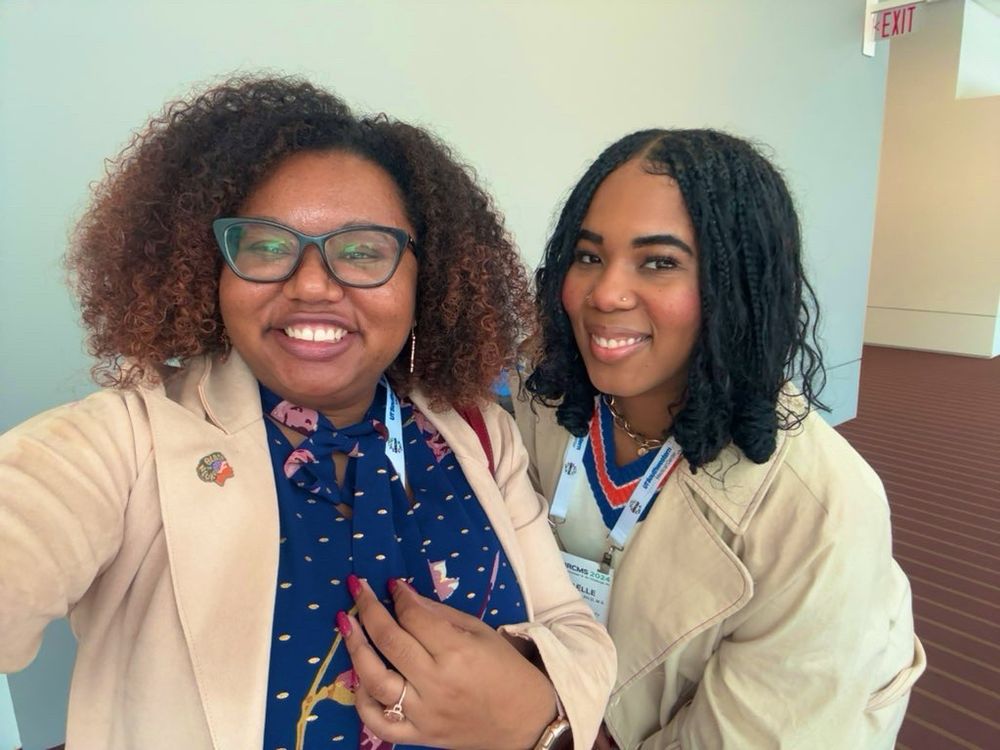 Two Haitian Black women PhDs taking a selfie. Dr. Dukes on the left has a curly Afro and is wearing a dark blue blouse with pink flowers and gold dots. She is also wearing a light pink blazer, Black In Neuro pin, and black glasses. Dr. Thomas on the right has a short braided bob and is wearing a brown sweater with a beige trench coat.