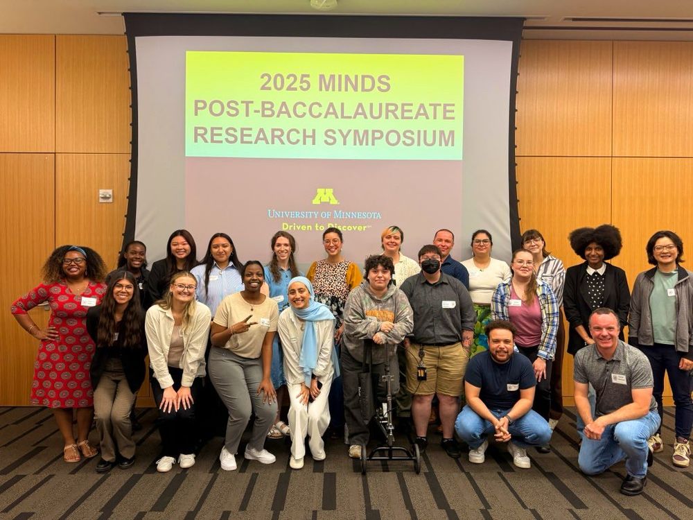 The 2025-2026 cohort of MINDS post-baccalaureate scholars, their near-peer graduate student mentors, faculty mentors, program directors, and mini-MINDS participants standing in front of a project screen that reads 2025 MINDS post-baccalaureate research symposium