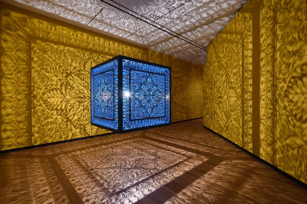 a sculptural installation in a yellow room, with light emitting from within a structure that casts ornate shadows onto the walls