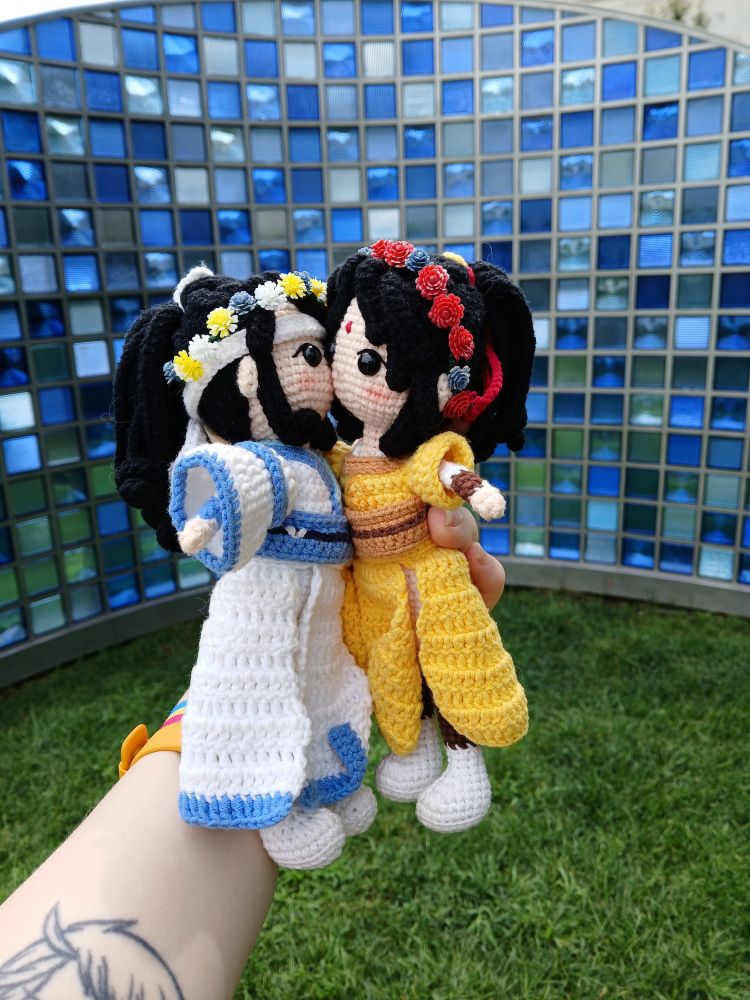 Crochet doll Lan Sizhui kissing the cheek of crochet doll Jin Ling in front of sculptural wall made up of glass squares in various shades of blue