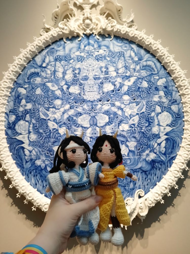 Crochet dolls of Lan Sizhui and Jin Ling held up in front of a large blue and white painted porcelain sculpture