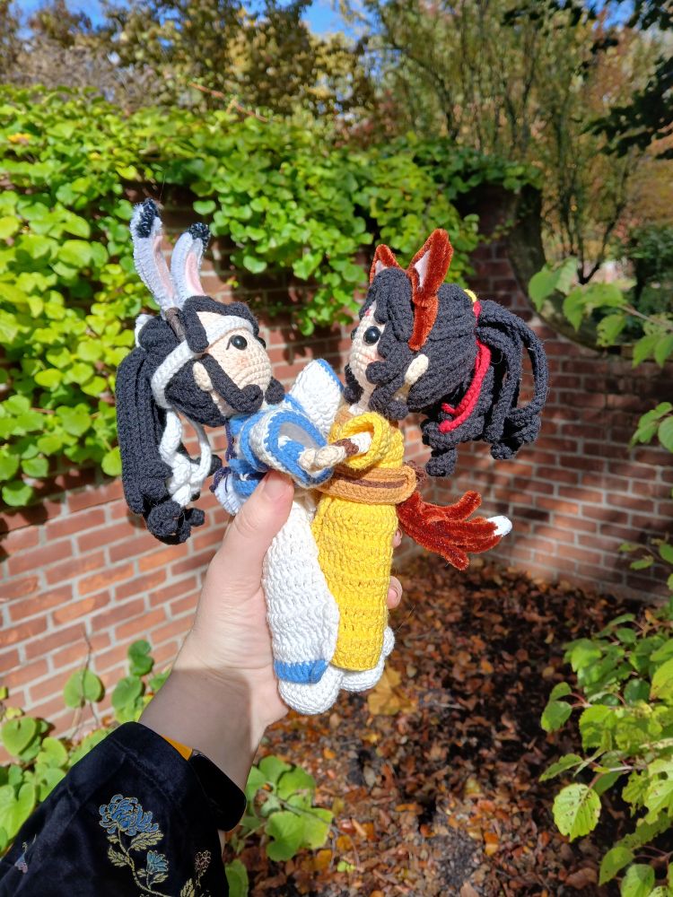 Side view of crochet dolls of Lan Sizhui with black-tipped rabbit ears seeming to hold hands with Jin Ling wearing brown dog ears and a tail