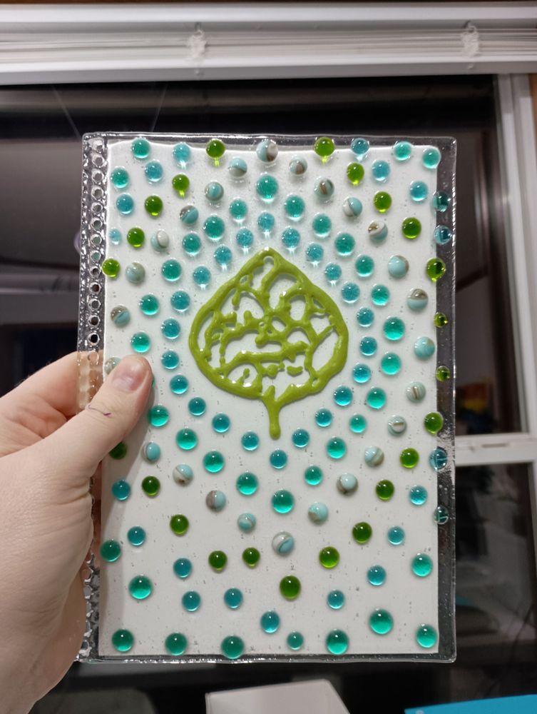 In front of a dark window, a caucasian hand holds up a transparent glass rectangle with a piece of white paper behind it. The paper is slightly smaller than the glass. The glass has a bunch of small holes in a line along the left side. The pattern on the glass is a leaf in the upper center, surrounded by dots in the color order: sky blue, blue-green, turquoise with white and brown, green, sky blue, blue-green. The lines of dots get increasingly unaligned and erratic the farther away from the leaf they get.