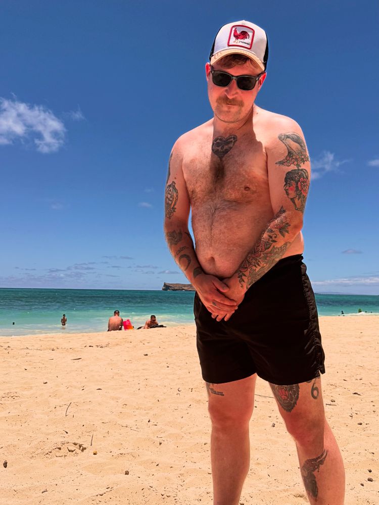 Me on the beach in Hawaii wearing black shorts and a hat with a red rooster on it.