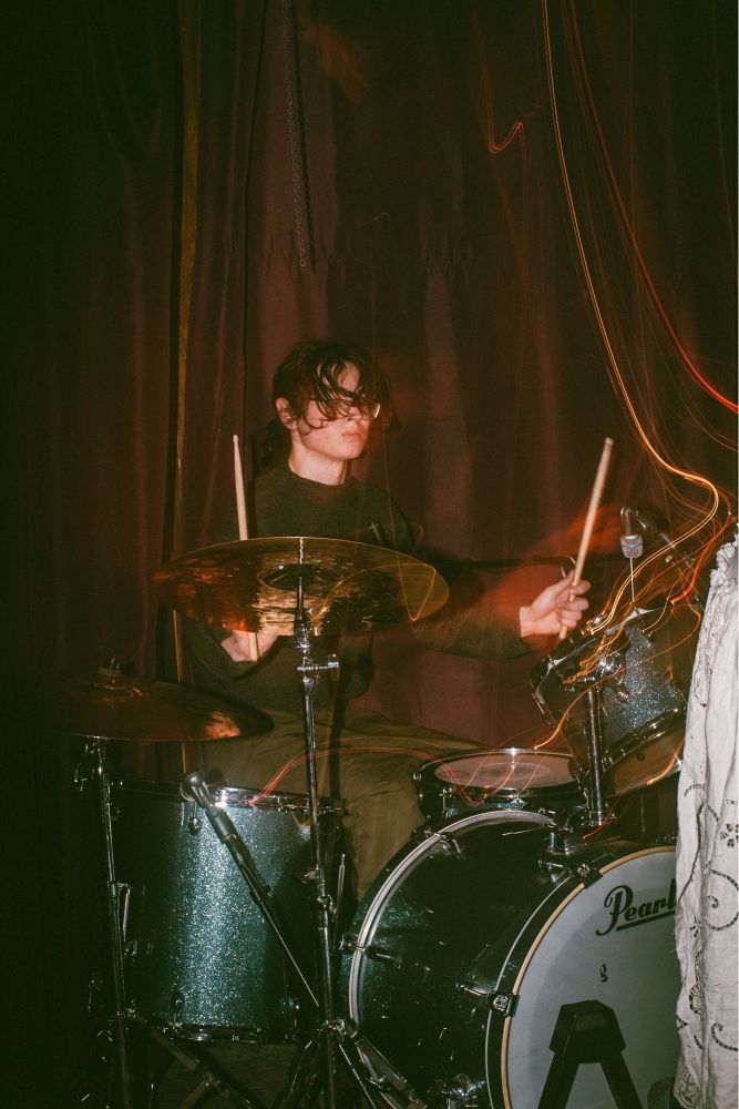 flash photography of siena, the drummer, at her drumkit. she is in a black sweater and a dark green skirt.