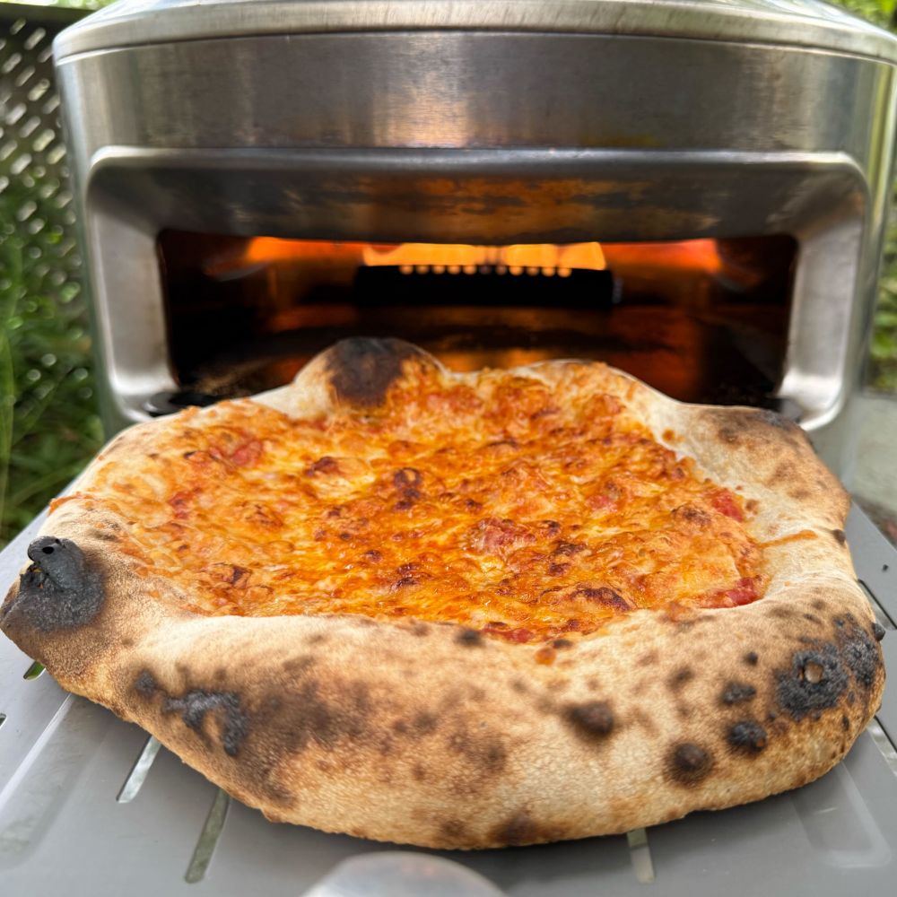 Cheese pizza on a pizza peel in front of a small pizza oven. The cheese has browned and there is some leoparding  on the crust. There are flames in the back of the stainless steel pizza oven.