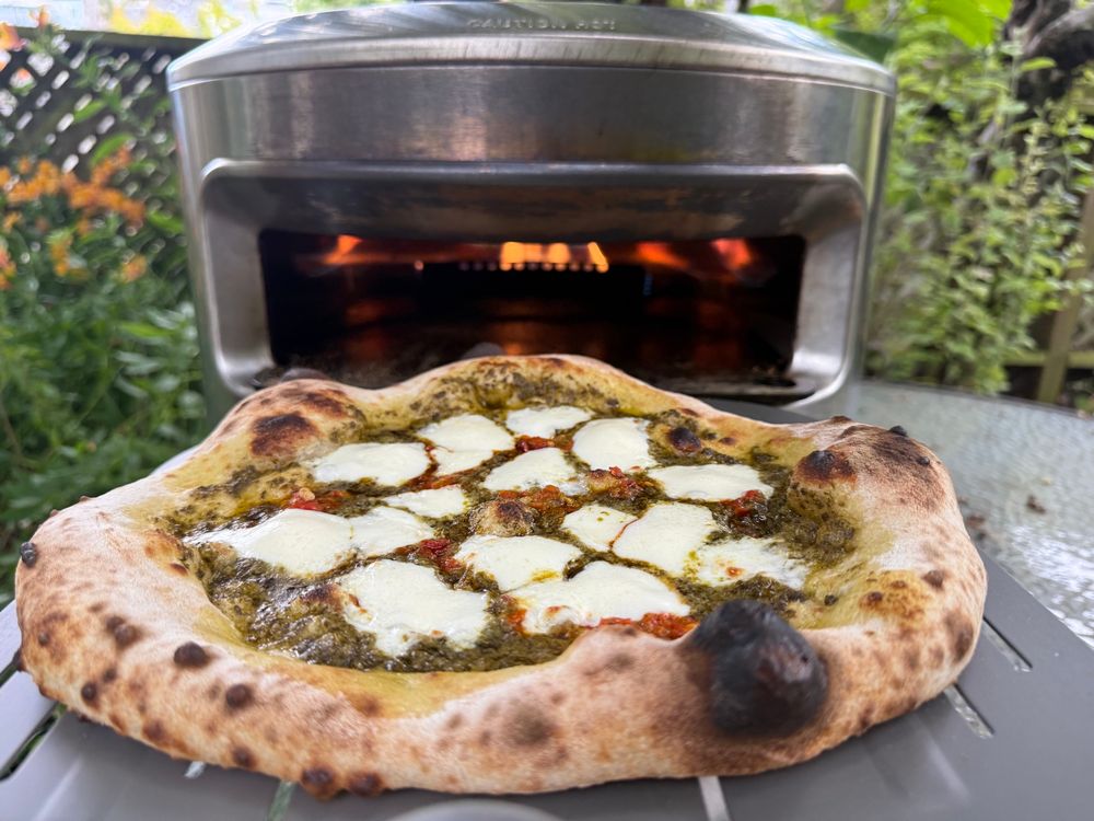 Pesto, Calabrian pepper, and fresh mozz pizza on a pizza peel coming out of a pizza oven 
