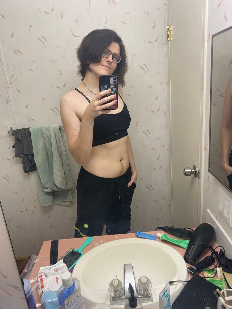 A futch lesbian in front of a mirror wearing a sports bra and and joggers. She is smirking and has messy hair. 