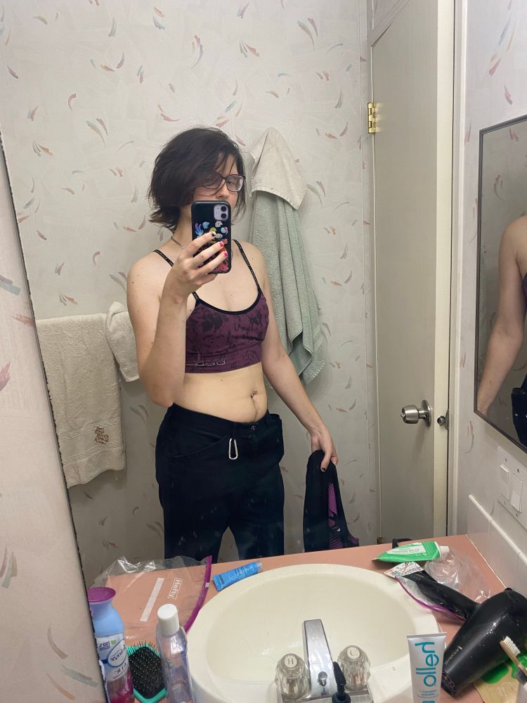 A lesbian trans girl with brown medium hair wearing black pants and a sports bra looks in a the mirror. She’s holding her longsleeve in one hand and a phone in the other. She has a carabiner