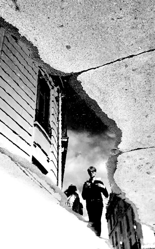 Un jeune homme de face croisant une personne de dos se reflètant dans une flaque d’eau. Noir et blanc.