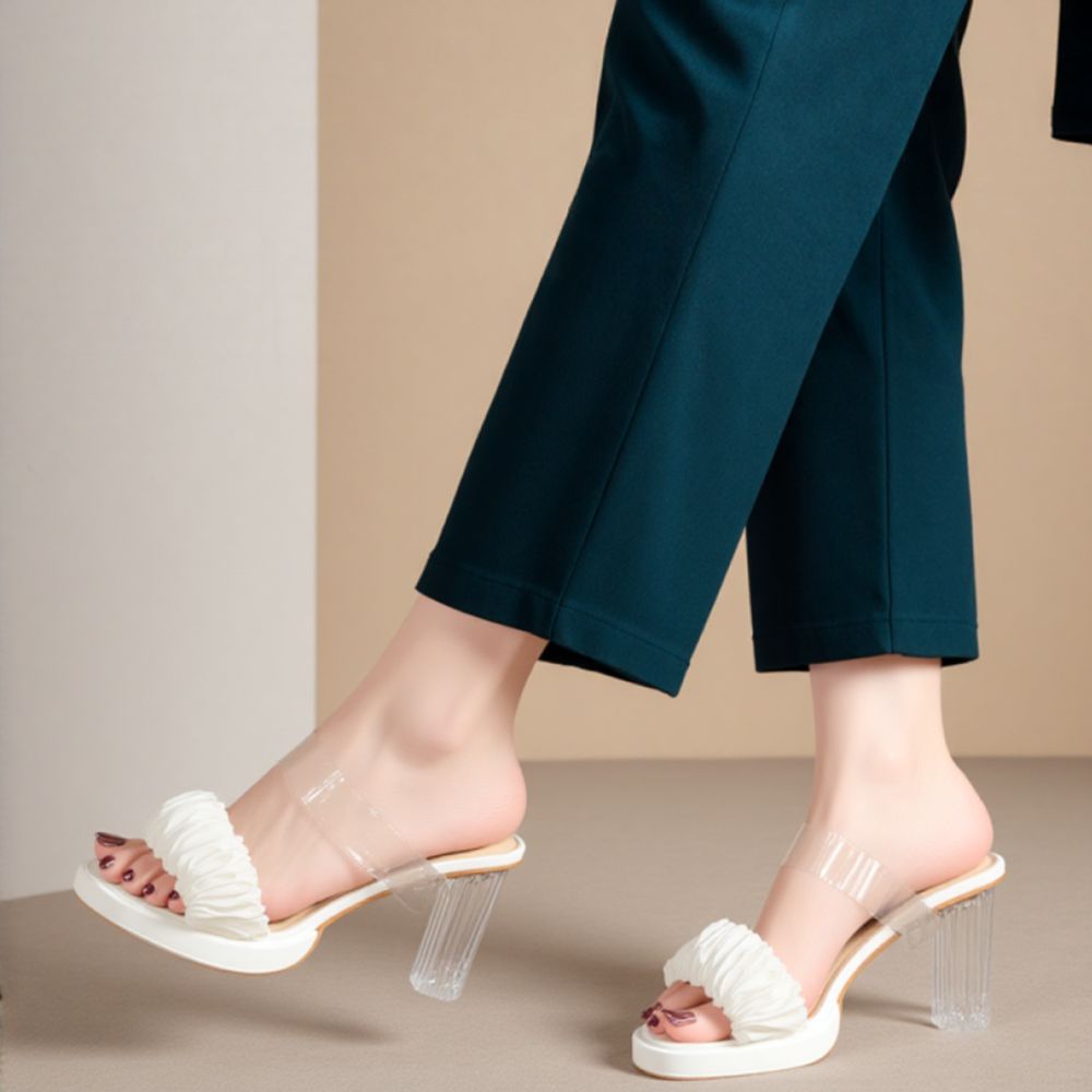 Close-up of white ruched strap, clear heels on model’s feet, minimalist aesthetic with neutral background — trendy summer shoes.

