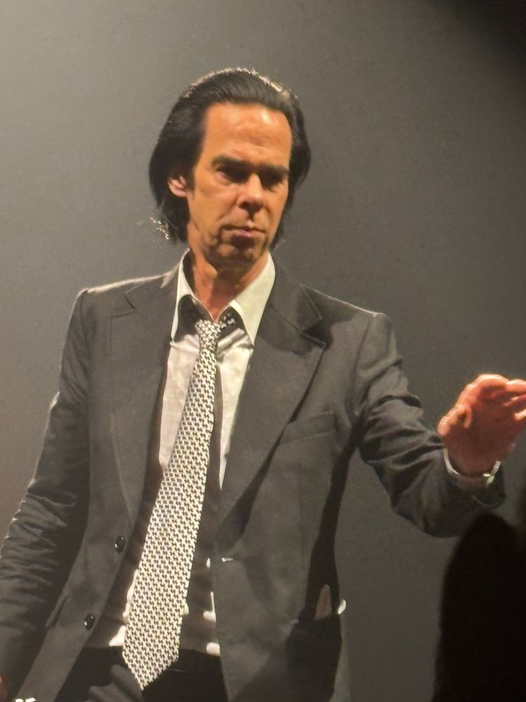a close up of nick cave on stage. he is looking over the crowd with his left hand extended