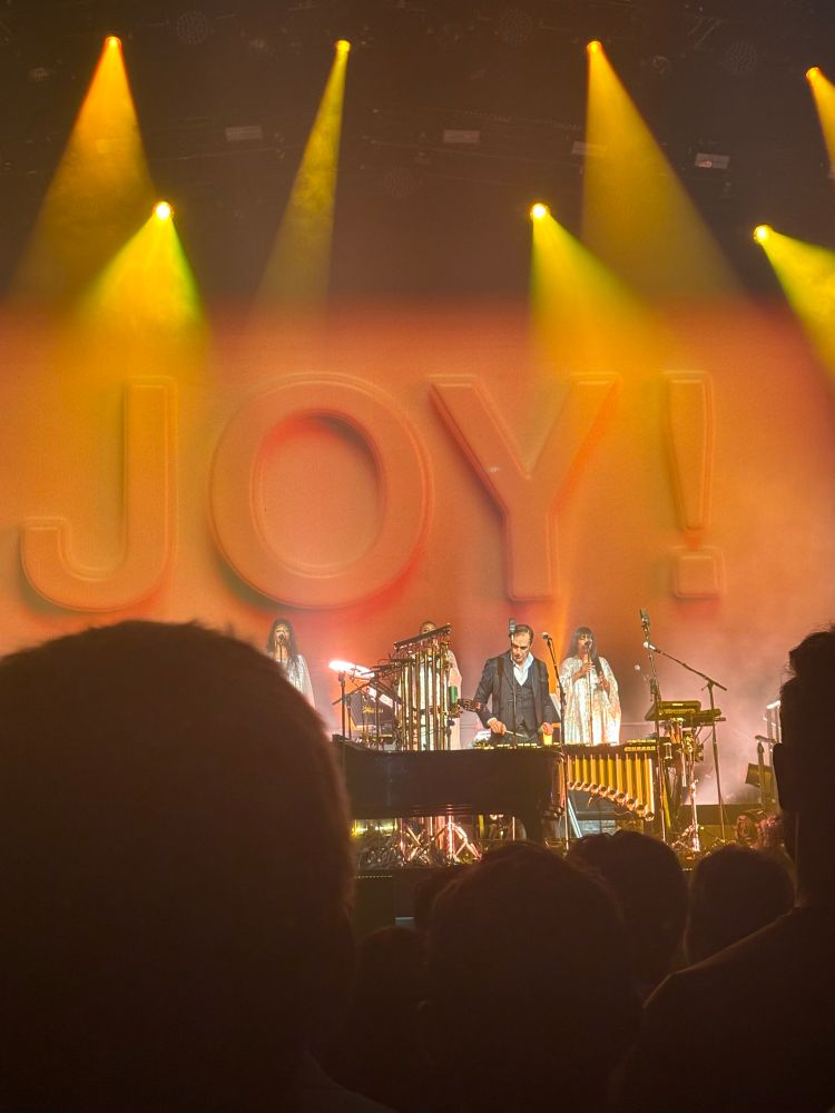 a shot of the stage during the song “joy”. the word “JOY!” is projected on the screen behind background gospel singers in red writing 