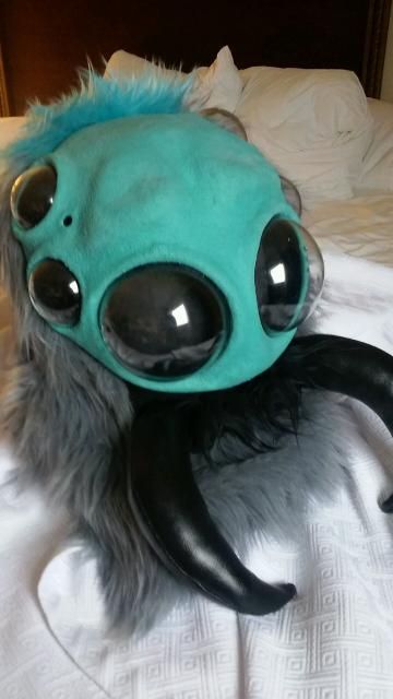 Jumping spider fursuit head with six large plastic eyes, black vinyl mandibles, teal skin, teal faux fur at the back, grey faux fur on the sides, and a little bit of black faux fur beneath the mandibles.