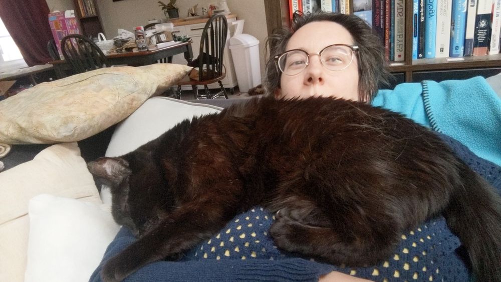 Me (a white human with short brown hair and glasses wearing a navy blue sweater) leaning way back on a couch piled with pillows and blankets. A sleeping black cat is draped fully across my chest from shoulder to shoulder. One of his front paws is reaching down to rest on my arm and the fur of his back is covering my mouth.