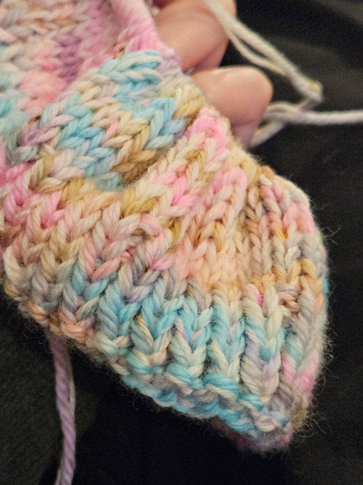 Close-up of in-progress knit featuring yarn that shifts between blue, pink, tan, beige, and lavender, giving the impression of cotton candy mixed with a root beer float.