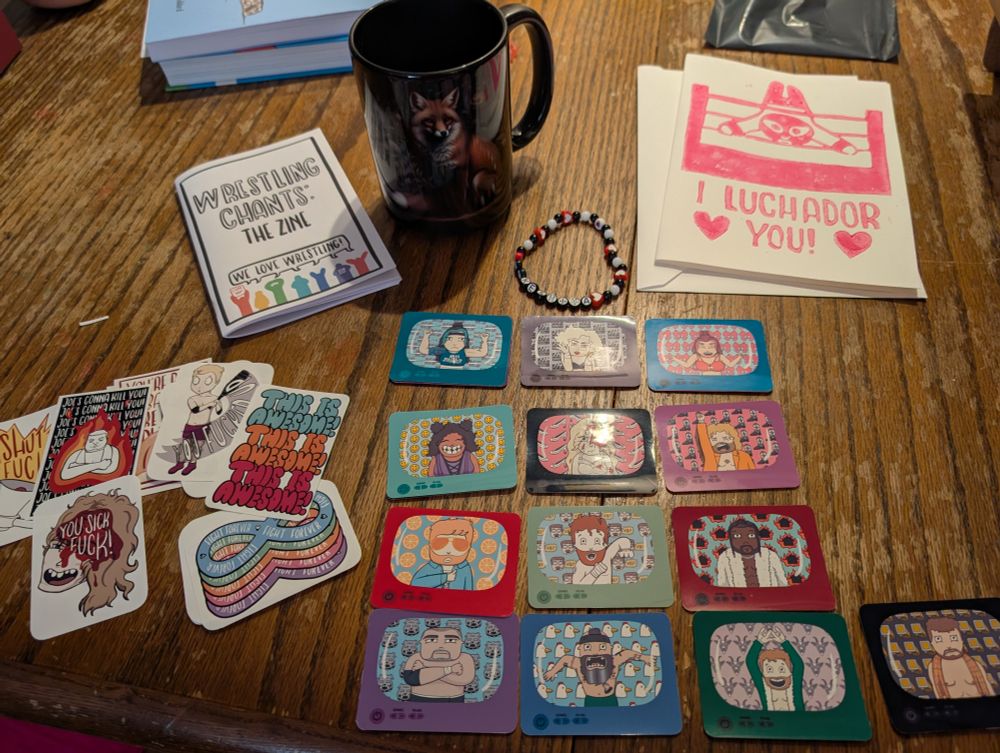On a wooden table: a wrestling chant zine and accompanying stickers, an 'i luchador you' card, a Takeshita bracelet, and cartoon TV stickers depicting Toni storm and lovers, the conglomeration and 'the new four pillars'. There's also a somehow gothic and chintzy big black mug with a fox on it.