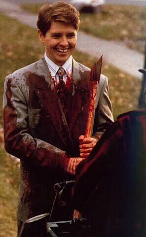 Still photo of a young Dave Foley from Kids in the Hall as an axe murderer. He is wearing a grey suit covered in blood, holding a bloody axe, and sporting a huge grin.