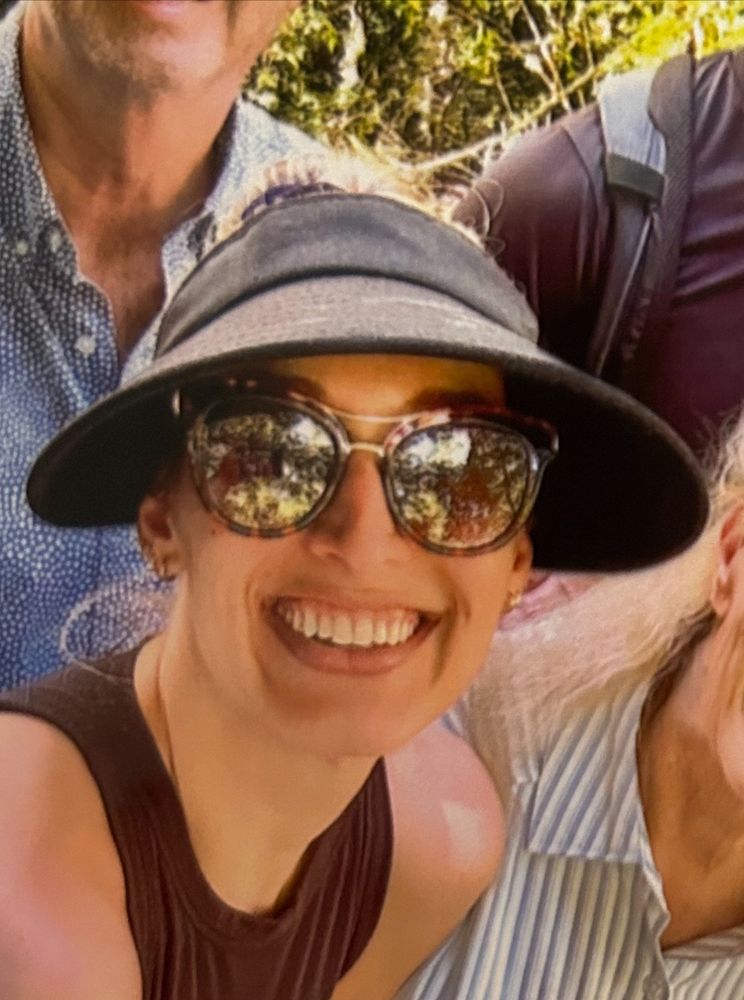 A close up photo of a young woman smiling broadly, wearing oversized sunglasses and a wide rimmed black hat. 